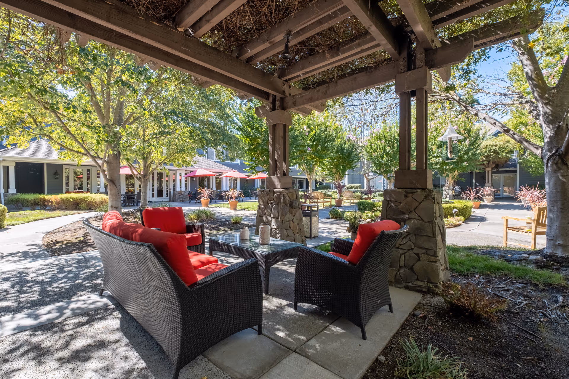 Outdoor seating area under a wooden pergola with stone pillars, featuring black wicker furniture with red cushions around a glass-top coffee table. The area is surrounded by trees, plants, and a paved walkway, with a building and red umbrellas visible in the background under a clear blue sky.