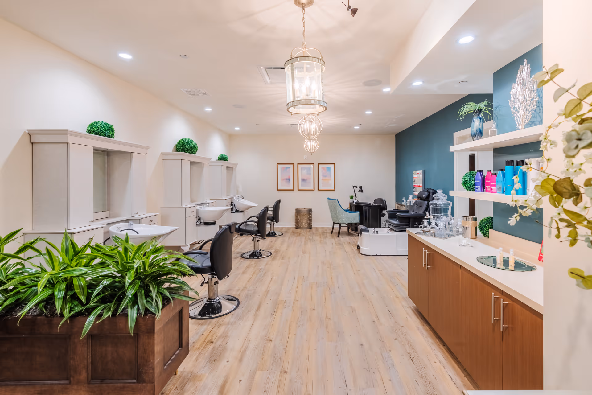 A bright and modern salon area in a senior living facility featuring multiple styling stations with chairs and sinks along the left wall, a wooden floor, decorative plants, shelves with hair care products on the right, and a pedicure chair with a small desk and chair in the back.