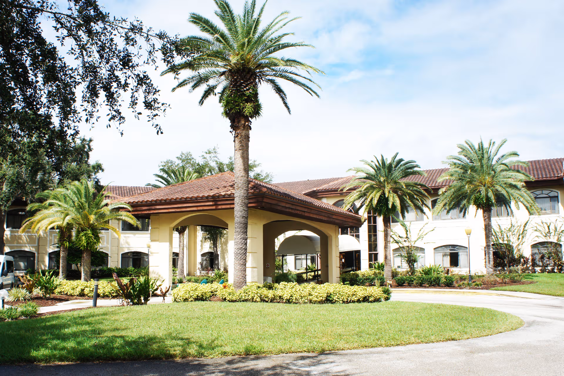 Exterior view of Westchester of Winter Park facility featuring a two-story building with a tiled roof, surrounded by palm trees and landscaped greenery under a partly cloudy sky.