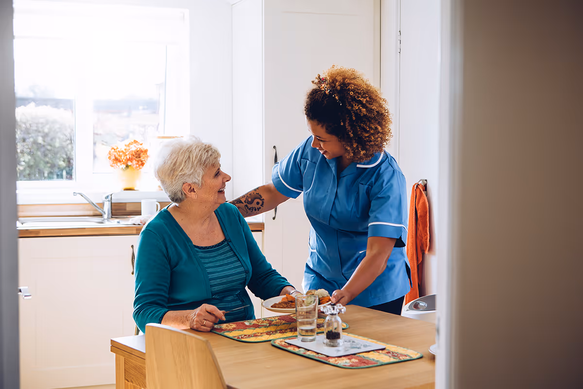 An elderly woman sitting at a kitchen table smiling at a caregiver in a blue uniform who is serving her a plate of food. The kitchen is bright with a window, a sink, and a vase with flowers in the background.