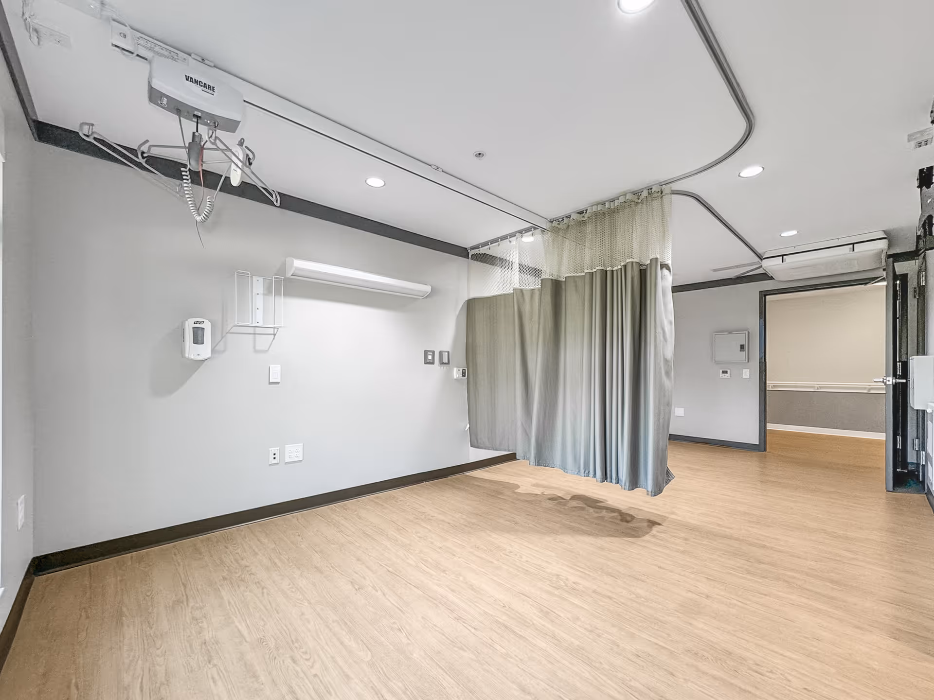 An empty medical room with light wood flooring, gray walls, and a ceiling-mounted patient lift system. A privacy curtain is partially drawn in the middle of the room. The room has recessed lighting and an open doorway leading to another area.