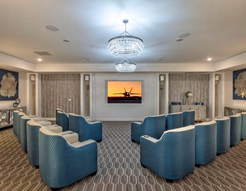 A modern media room with two rows of blue cushioned chairs facing a wall-mounted flat screen TV displaying an aircraft at sunset. The room features patterned carpet, two crystal chandeliers, decorative wall art, and side cabinets with vases and decor items.