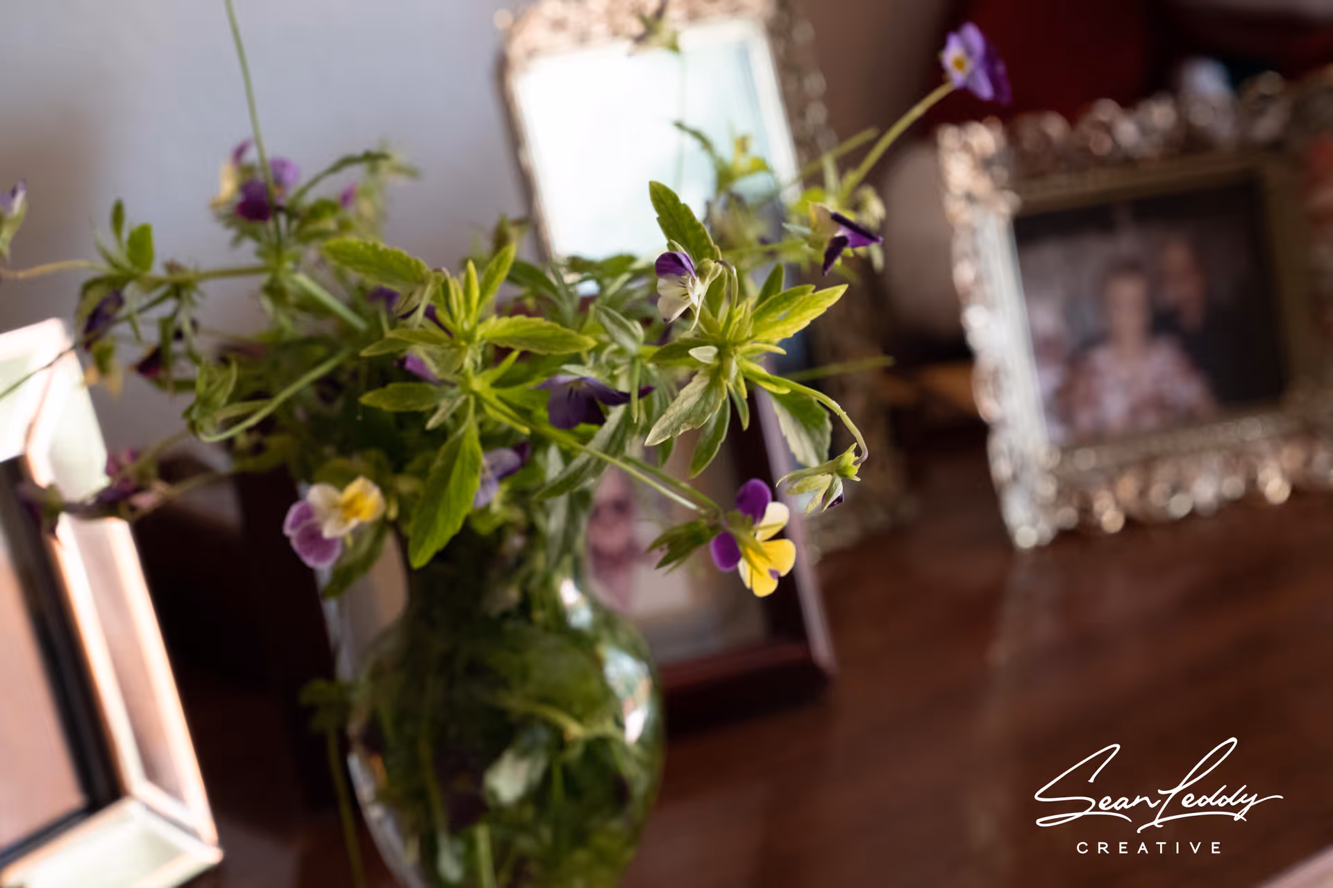 A close-up of a glass vase with green leaves and small purple and yellow flowers on a wooden surface, with blurred framed photographs in the background.