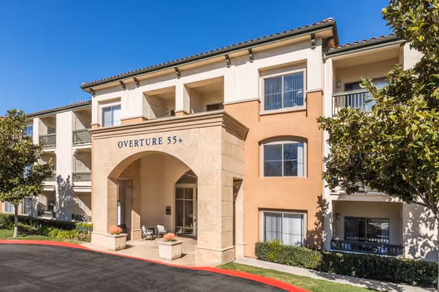Exterior view of a three-story senior living facility building with a beige and light brown facade, featuring a large archway entrance labeled 'OVERTURE 55+'. There are windows with blinds, small shrubs, and trees around the building, and a paved driveway with a red curb in front.