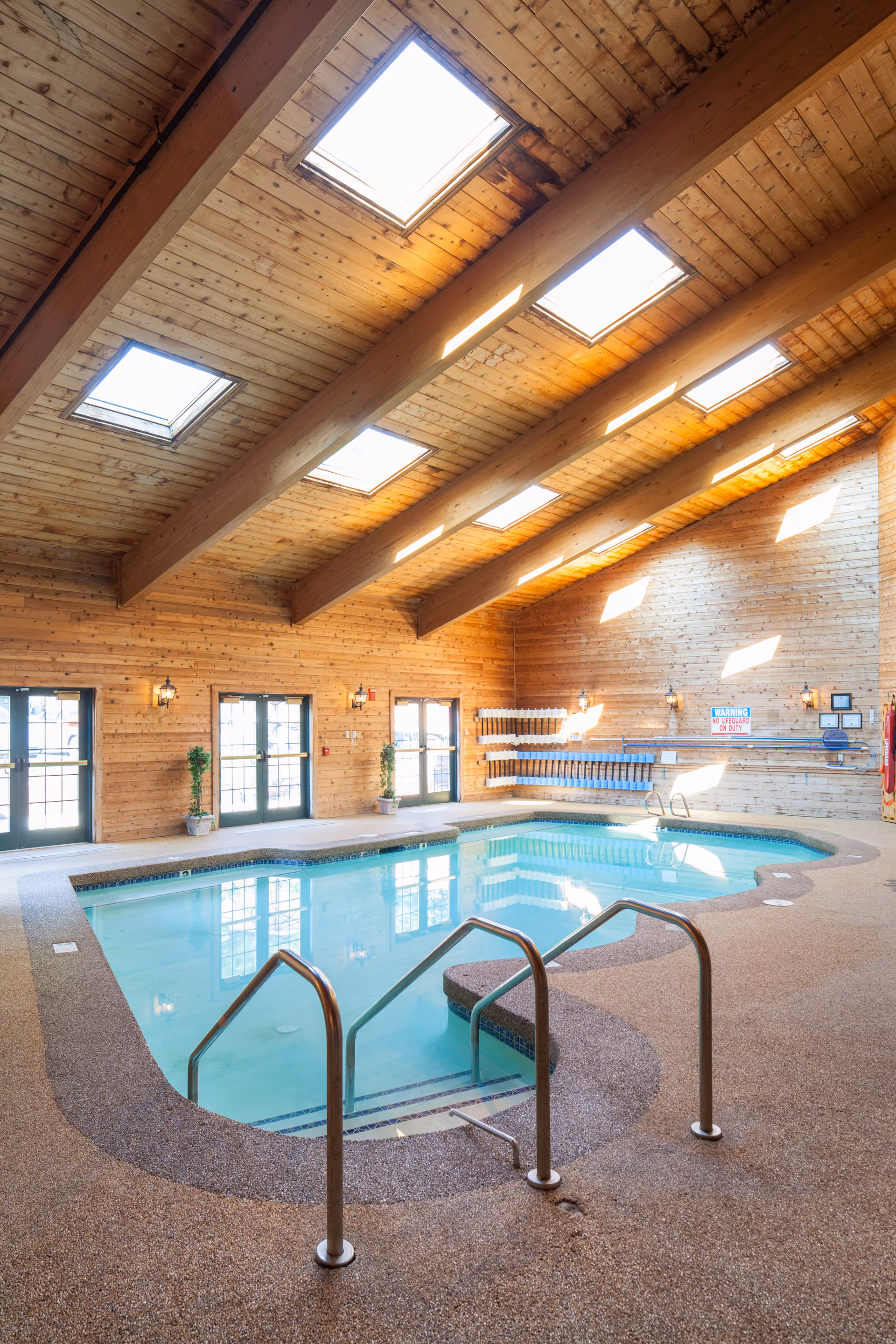 Indoor swimming pool with clear blue water inside a wooden room featuring a high ceiling with skylights. The pool area has metal handrails, textured flooring, and several doors and windows along the walls letting in natural light. There are also potted plants and pool equipment mounted on the walls.