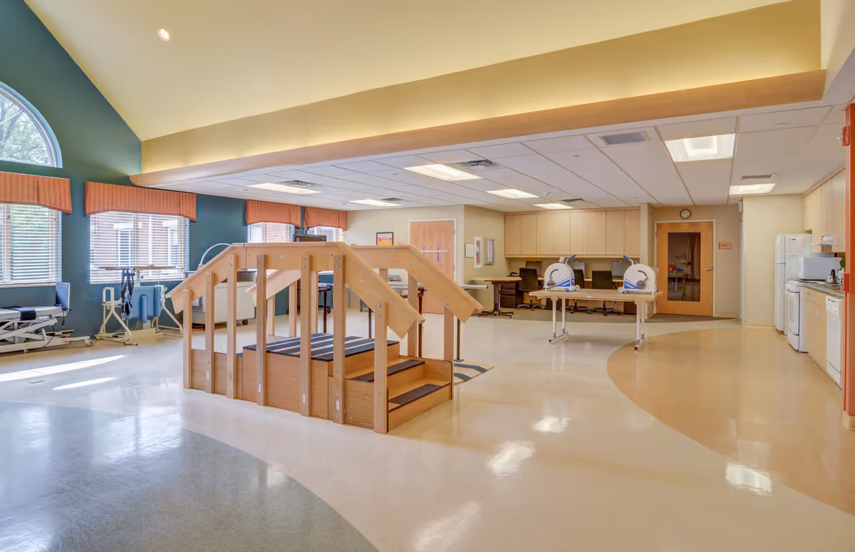 A spacious rehabilitation room with a wooden exercise staircase in the center, medical equipment along the walls, large windows with orange valances, and a kitchenette area with white appliances on the right side.