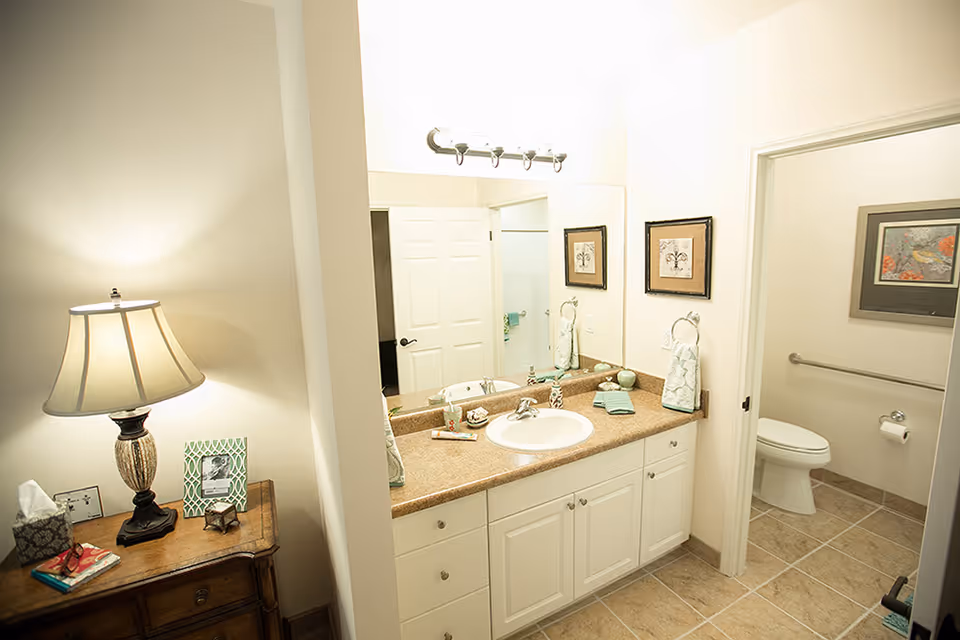 A well-lit bathroom featuring a countertop with a sink, hand towels, and decorative items. To the right, there is a separate toilet area with a grab bar and framed artwork on the wall. On the left side, a wooden table holds a lamp, tissue box, picture frame, and some books.