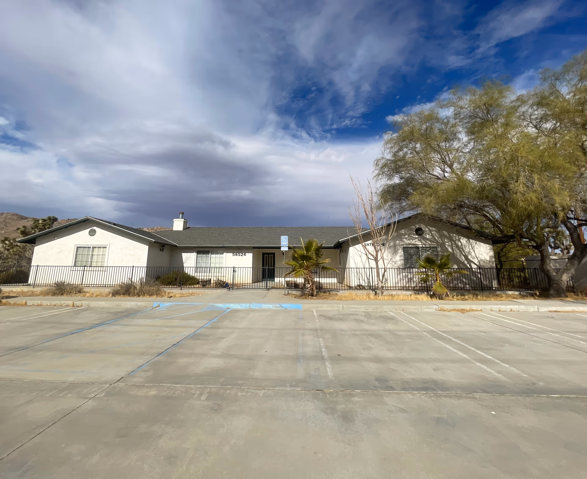 Single-story building with a light exterior, green roof, and several windows, surrounded by a black metal fence. There are a few small palm trees and a large tree on the right side. The foreground shows an empty concrete parking lot with marked handicap parking spaces. The sky above is partly cloudy with patches of blue.