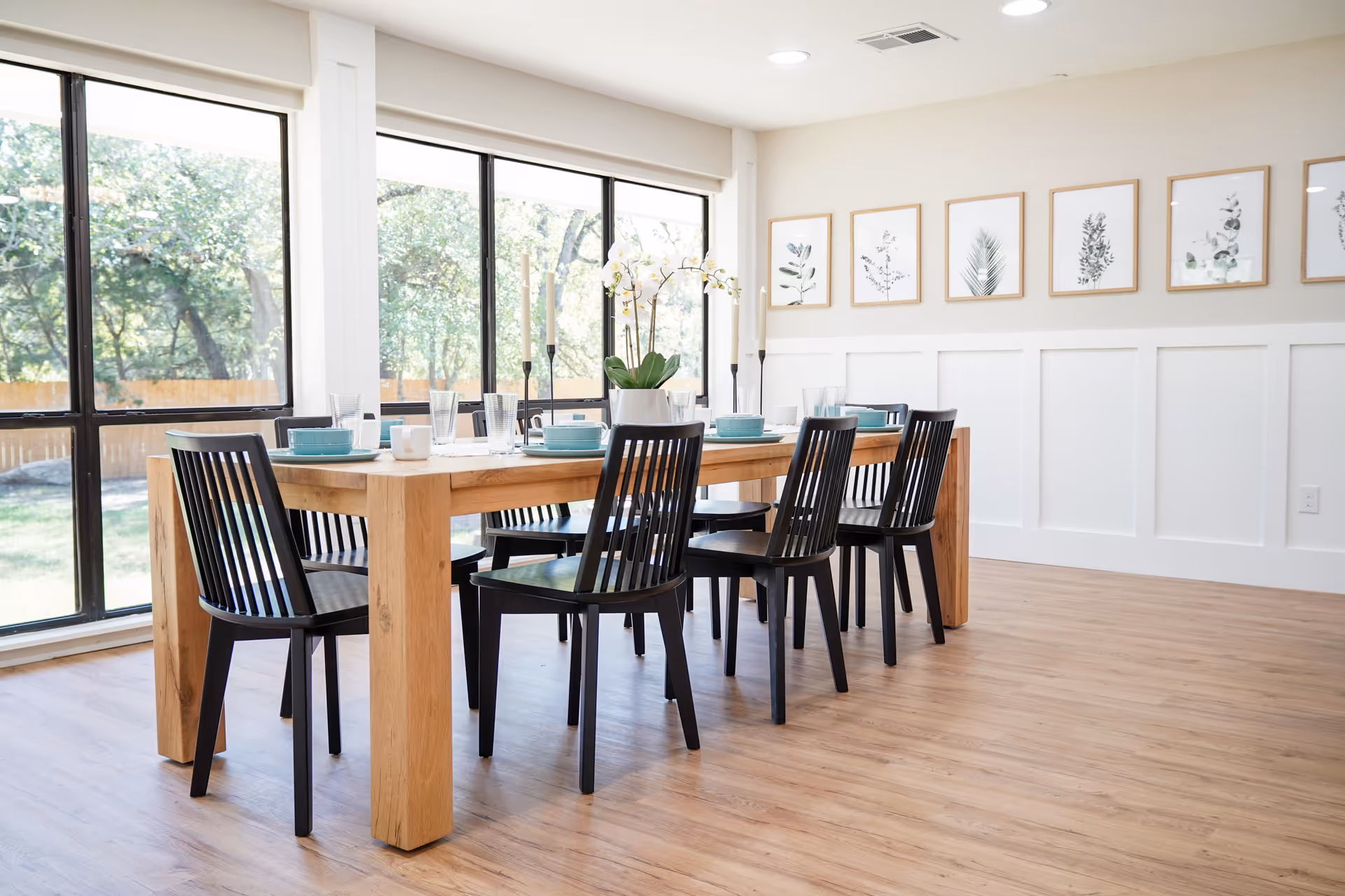 Bright dining room with a large wooden table set for six, black chairs, and large windows overlooking trees.