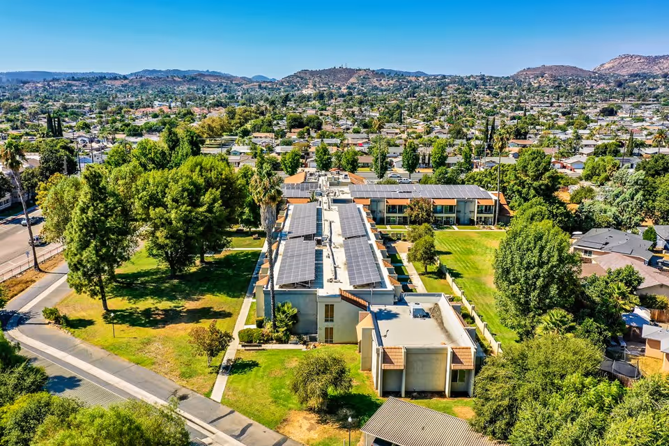 Aerial view of a senior living facility with multiple buildings featuring solar panels on the roofs, surrounded by green lawns, trees, and a suburban neighborhood with hills in the background.