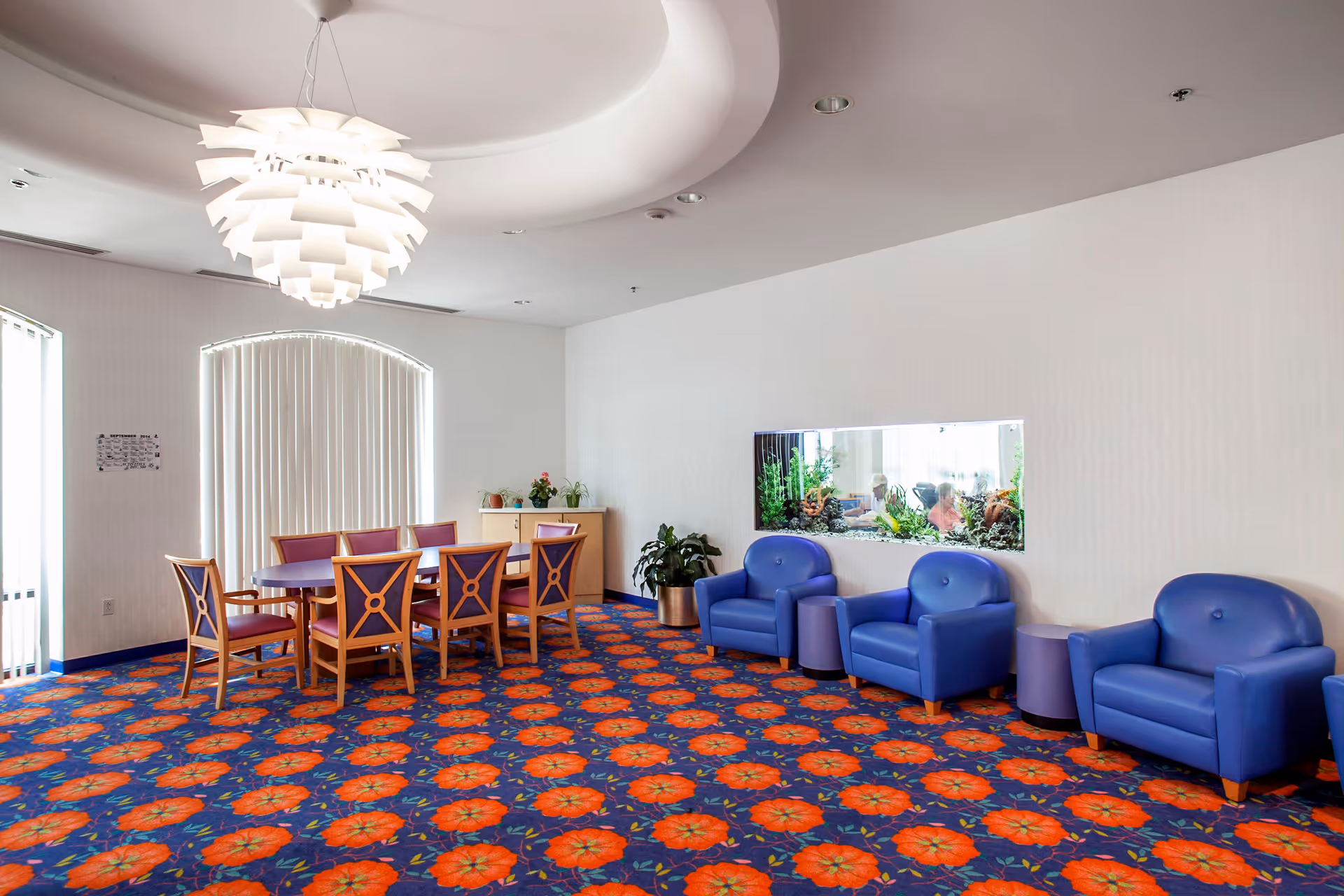 A brightly lit room with a colorful carpet featuring an orange floral pattern on a blue background. The room has a wooden dining table with eight chairs, each with purple cushions. On the right side, there are three blue armchairs separated by small purple tables. A large rectangular fish tank is embedded in the white wall behind the armchairs. The ceiling has a modern white pendant light fixture and a circular recessed design. There are vertical blinds covering the windows on the left side, and a small cabinet with plants on top is positioned against the back wall.
