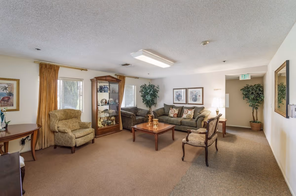 A cozy living room with beige carpet and white walls, featuring a green sofa with decorative pillows, a matching loveseat, an armchair, and a wooden coffee table with decorative items. There is a glass display cabinet with various ornaments, two windows with golden curtains, framed artwork on the walls, a potted plant in the corner, and a hallway with an exit sign.