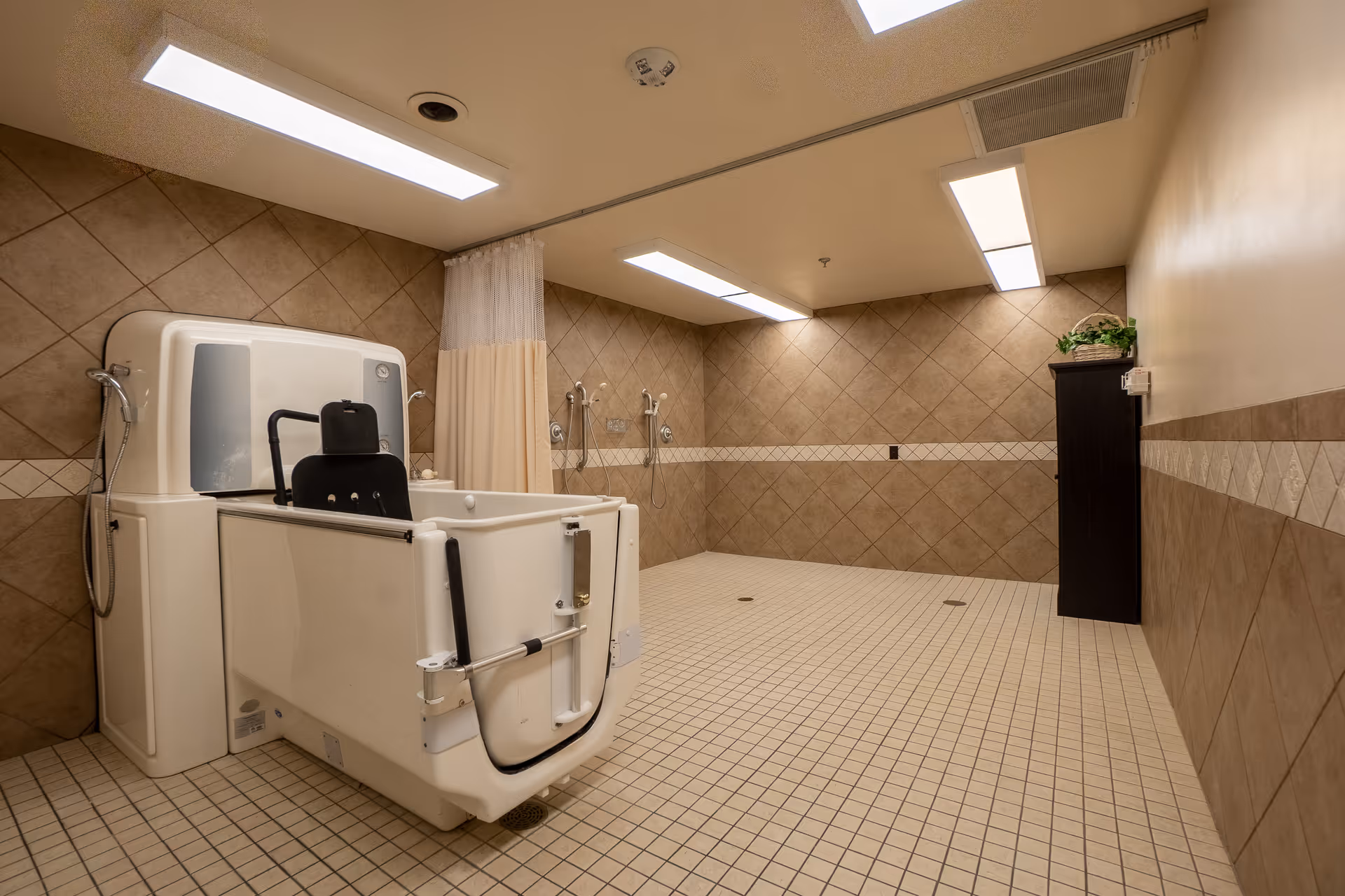 A spacious assisted bathing room with beige tiled walls and floor. The room features a large walk-in bathtub with a black seat and handrails. There are two shower heads mounted on the wall with a beige privacy curtain between them. The ceiling has fluorescent lighting, and a small dark cabinet with a plant on top is positioned against the far wall.
