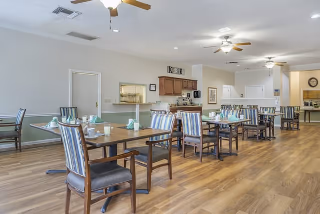 Spacious senior living dining room with multiple set tables and chairs, wood-look floors, ceiling fans, and a service counter.