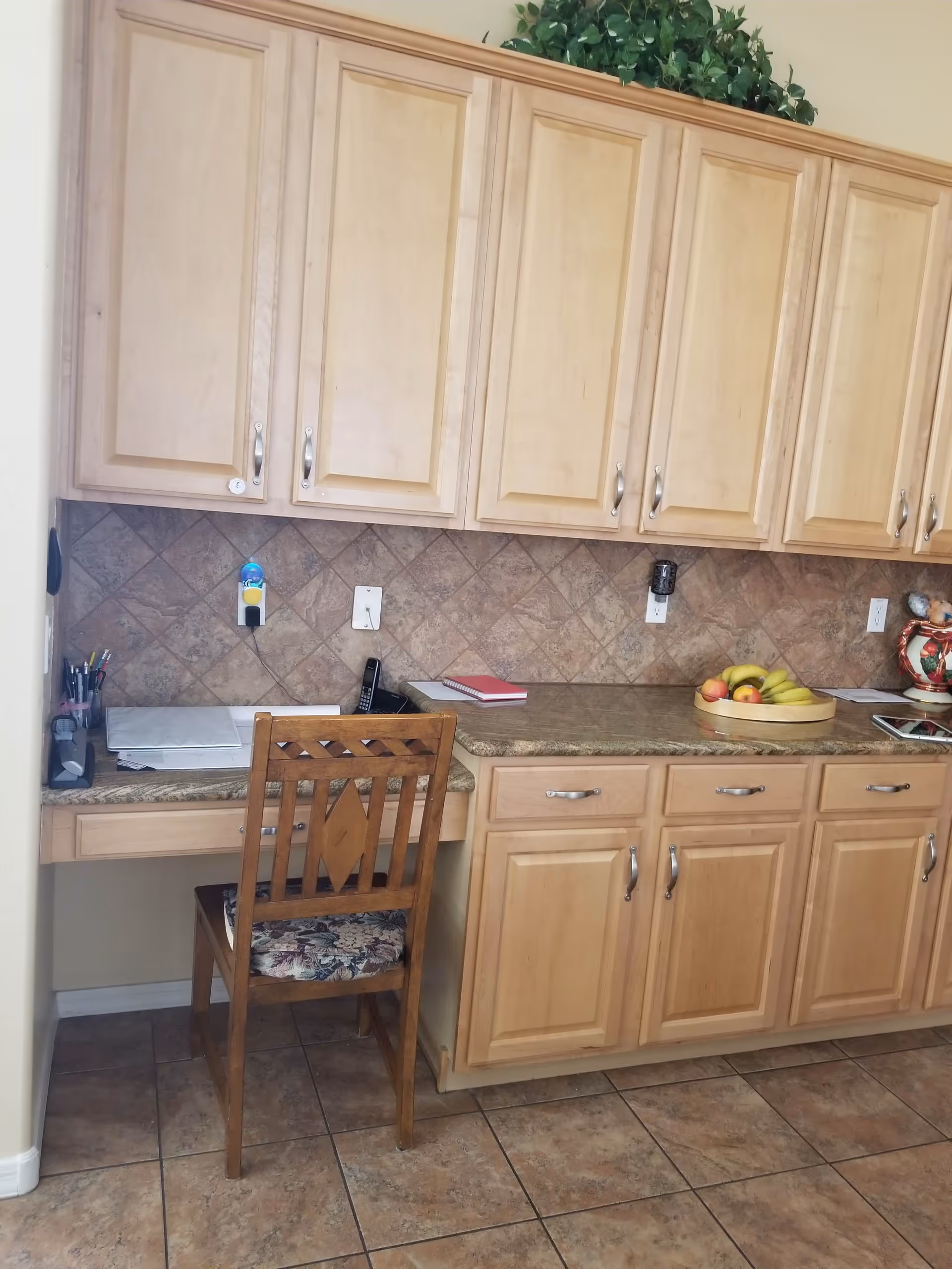 A kitchen area with light wood cabinets and a tiled backsplash. There is a wooden chair with a floral cushion in front of a small built-in desk space. On the countertop, there is a tray with bananas and apples, a notebook, a cordless phone, and some office supplies. The floor is tiled with brown tiles.