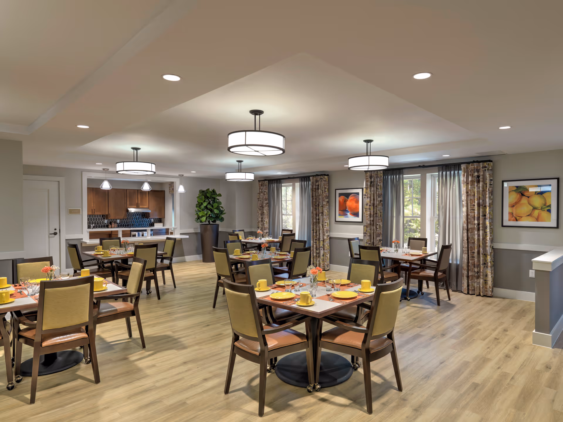 A bright and spacious dining room with several square wooden tables set for meals, each with yellow cups, plates, and orange flowers in small vases. The room has light wood flooring, neutral-colored walls, large windows with patterned curtains, and modern ceiling lights. In the background, there is a kitchen area with wooden cabinets and a tiled backsplash.