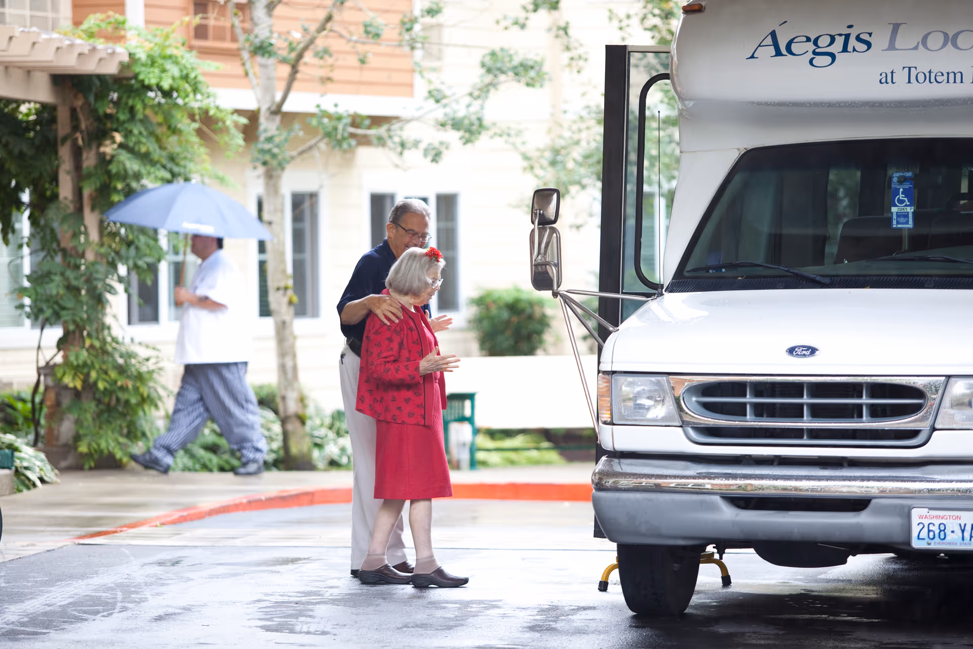 An elderly woman in a red dress and jacket is being assisted by a man as they stand near a white Aegis Lodge shuttle bus. In the background, a person holding a blue umbrella walks past a building with windows and greenery.