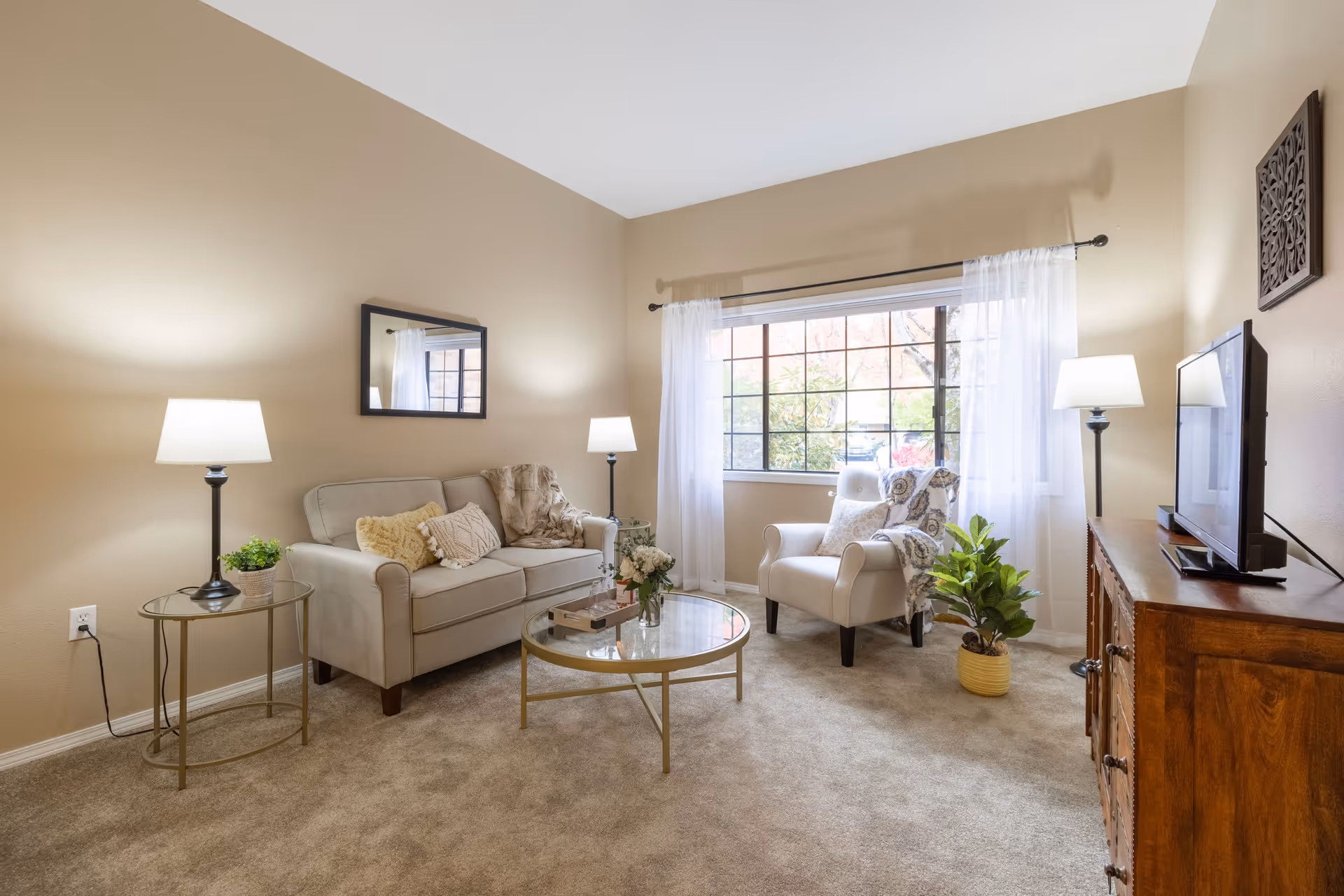 A cozy living room with beige walls and carpet. The room features a beige sofa with decorative pillows and a throw blanket, a white armchair with a patterned throw, a round glass coffee table with flowers and books, two side tables with lamps, a wooden TV stand with a flat-screen TV, and a large window with sheer white curtains letting in natural light. There are also two potted plants and a wall mirror above the sofa.