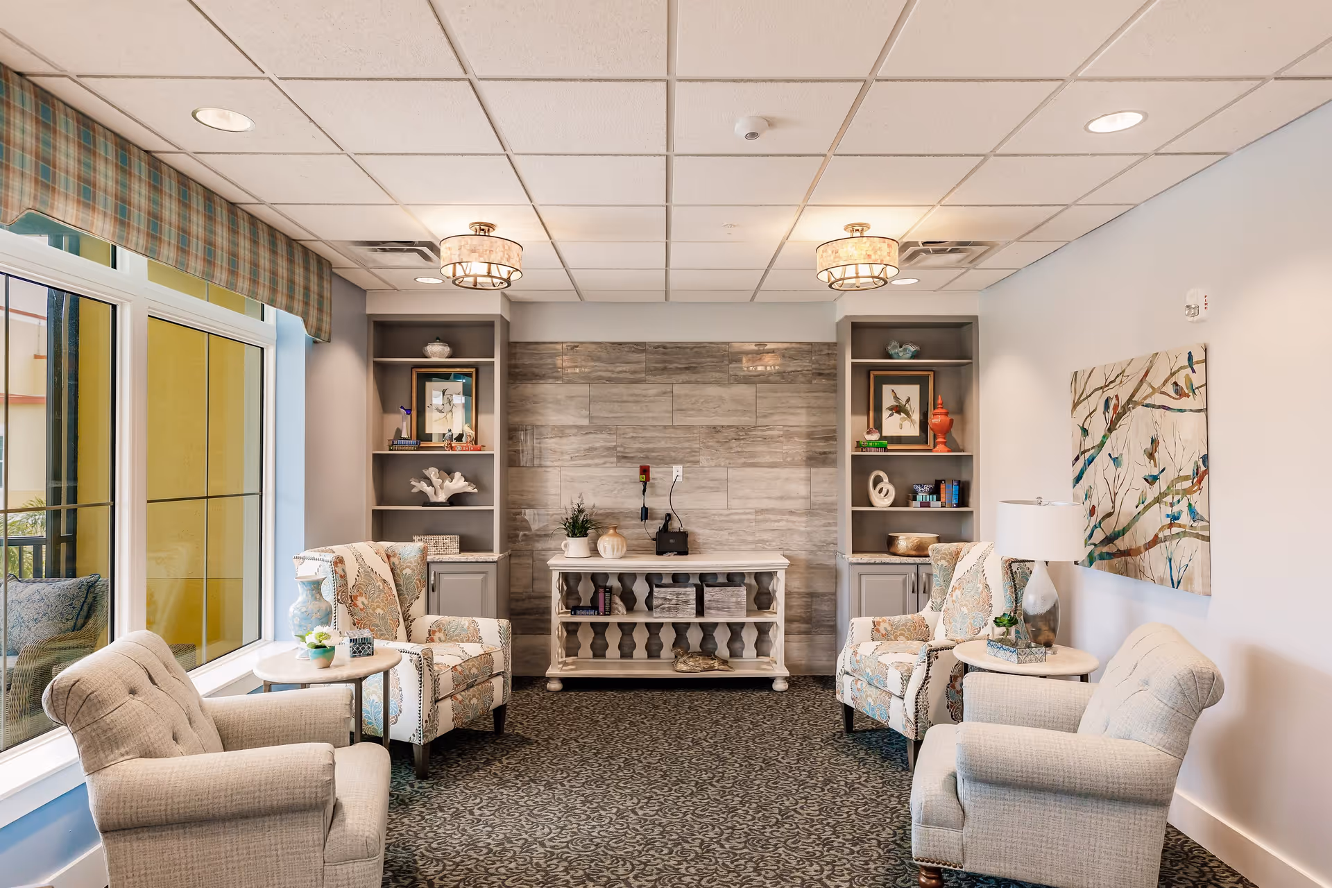 A cozy sitting area in a senior living facility with four upholstered armchairs arranged around two small round tables. The room features built-in shelves with decorative items, a console table against a tiled accent wall, and large windows with plaid valances letting in natural light. A painting of birds on branches hangs on one wall, and ceiling lights provide additional illumination.