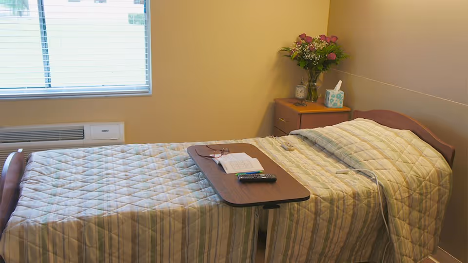 A single bed in a nursing facility room with an overbed table holding glasses, a remote and papers, and a bedside table with flowers and tissues.