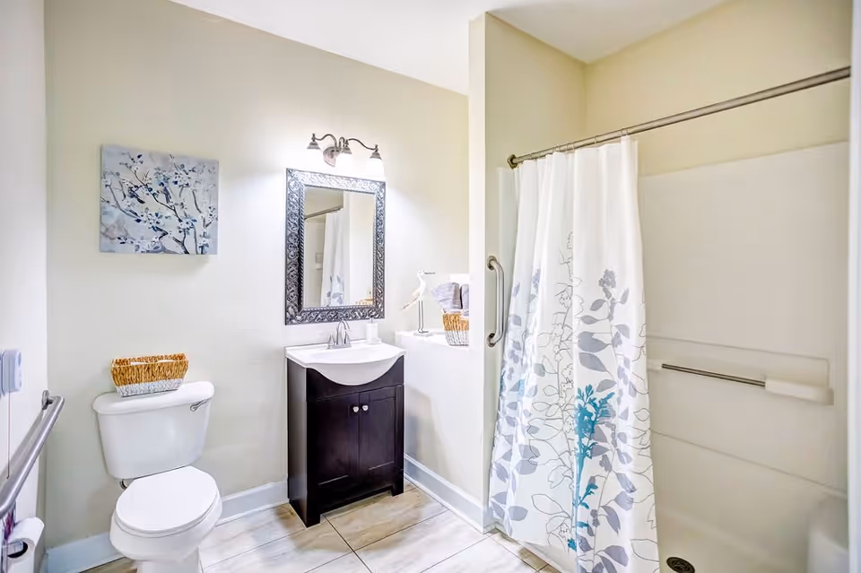 A clean and bright bathroom featuring a white toilet with a wicker basket on top, a dark wood vanity with a white sink, a decorative framed mirror above the sink, a wall-mounted light fixture, a shower with a white curtain decorated with blue and gray floral patterns, and a piece of wall art with a floral design.