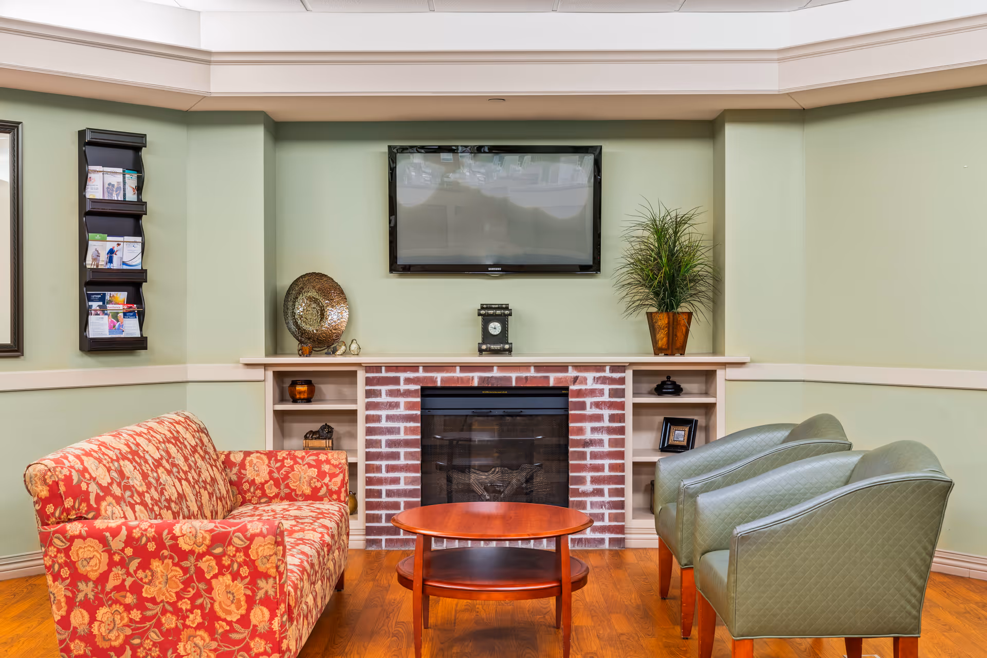 A cozy living room area with a red floral patterned sofa on the left, two green armchairs on the right, and a wooden coffee table in the center. The back wall features a brick fireplace with built-in shelves on either side, decorated with various items including a plant, a decorative plate, and framed pictures. Above the fireplace is a mounted flat-screen TV. The walls are painted light green with beige trim, and the floor is wooden.