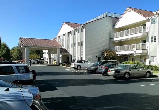 Exterior view of a multi-story senior living facility named Riverview Terrace with a covered entrance and a parking lot with several parked cars under a clear blue sky.