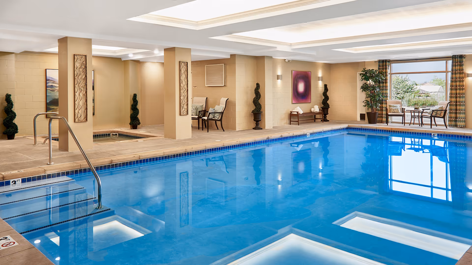 Indoor swimming pool area with seating, potted plants, and skylights in a senior living facility.
