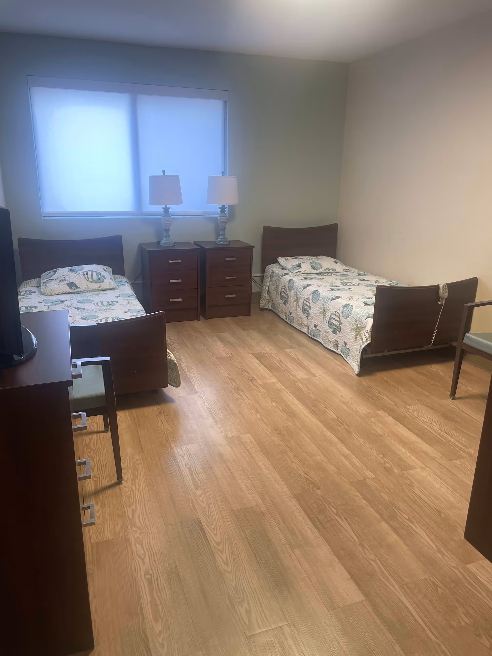 A simple bedroom with two single beds, each with a seashell-patterned quilt. Between the beds are two wooden nightstands with matching lamps. The room has light-colored walls, a large window with a closed blind, and wood-look flooring. There is a wooden dresser with a chair in the foreground.