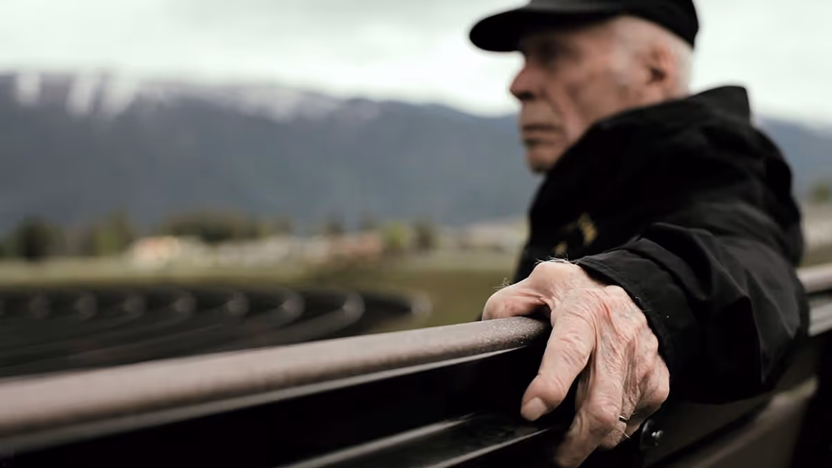 An elderly person wearing a black jacket and cap is sitting on a bench outdoors, with their hand resting on the back of the bench. The background shows blurred mountains and greenery.