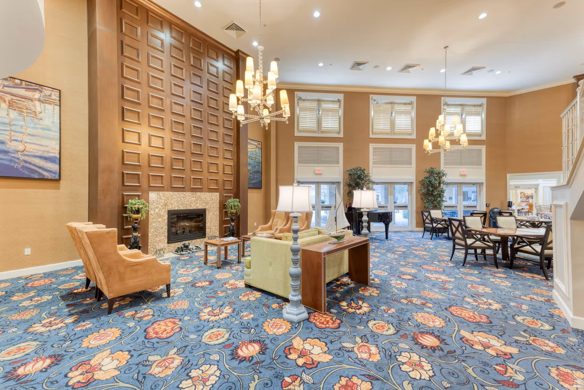 A spacious senior living facility common area with a high ceiling, large windows with shutters, and a patterned blue carpet with floral designs. The room features a wooden paneled wall with a fireplace, two beige armchairs, a green sofa with a wooden console table behind it, two standing lamps, a grand piano, and several dining tables with chairs. There are also two large plants near the windows and two chandeliers hanging from the ceiling.