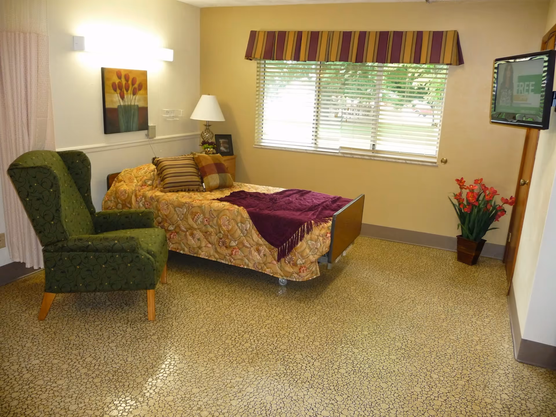 A small furnished bedroom with a single bed, green armchair, side table and lamp, a window with blinds, and a wall-mounted TV.