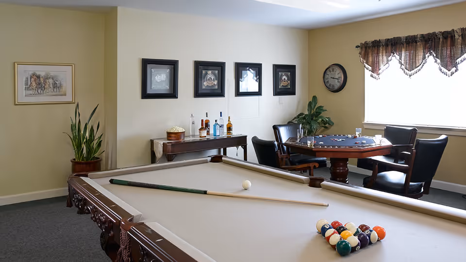 A recreational room with a beige pool table in the foreground, a pool cue and balls arranged for a game. In the background, there is a wooden table with bottles and a bowl of popcorn, four black leather chairs around a card table with playing cards and poker chips, a wall clock, framed pictures on the wall, a potted plant, and a window with a valance curtain.