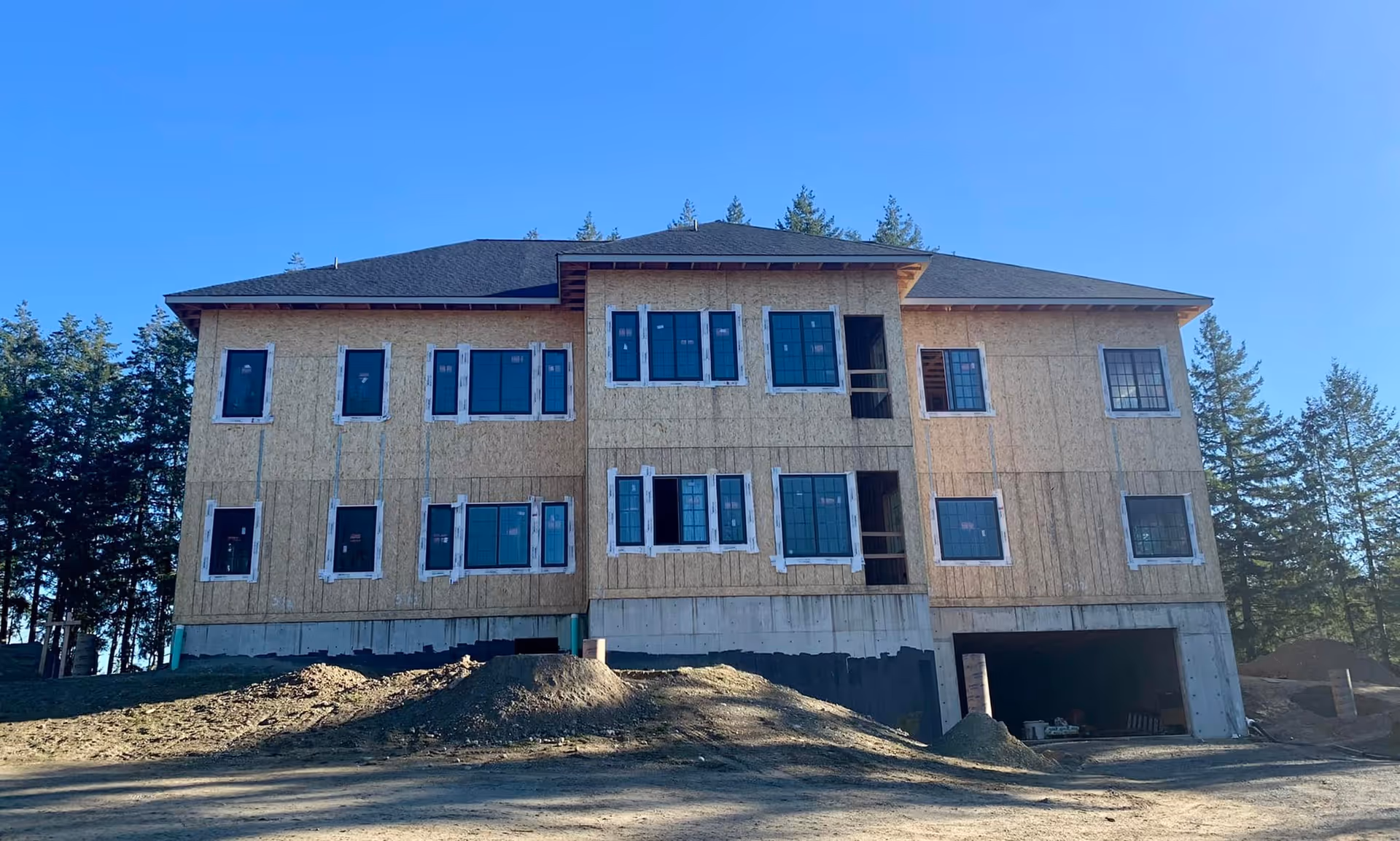 A large two-story building under construction with exposed wooden framing and multiple windows installed. The building sits on a concrete foundation with an open garage area on the right side. There are piles of dirt in front of the building and trees in the background under a clear blue sky.