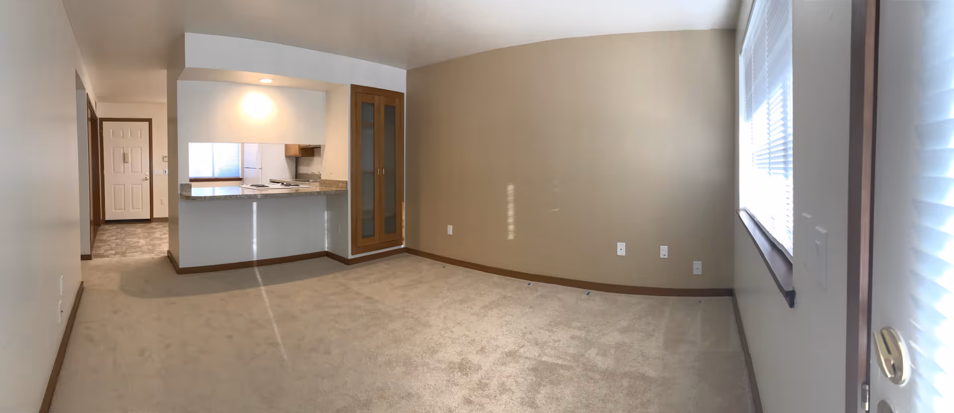Empty interior room with beige carpet and walls, a window with blinds on the right, a small kitchen area with a granite countertop and wooden cabinets in the background, and a door leading to another room on the left.