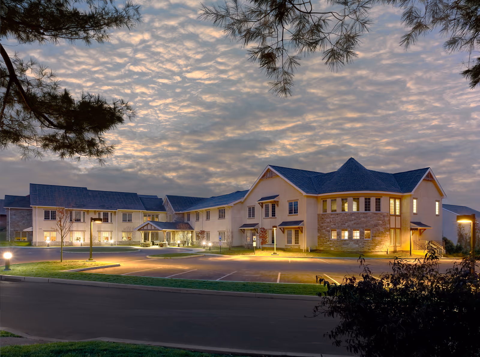 Front exterior of a two-story senior living facility at dusk with illuminated windows and a landscaped parking area.