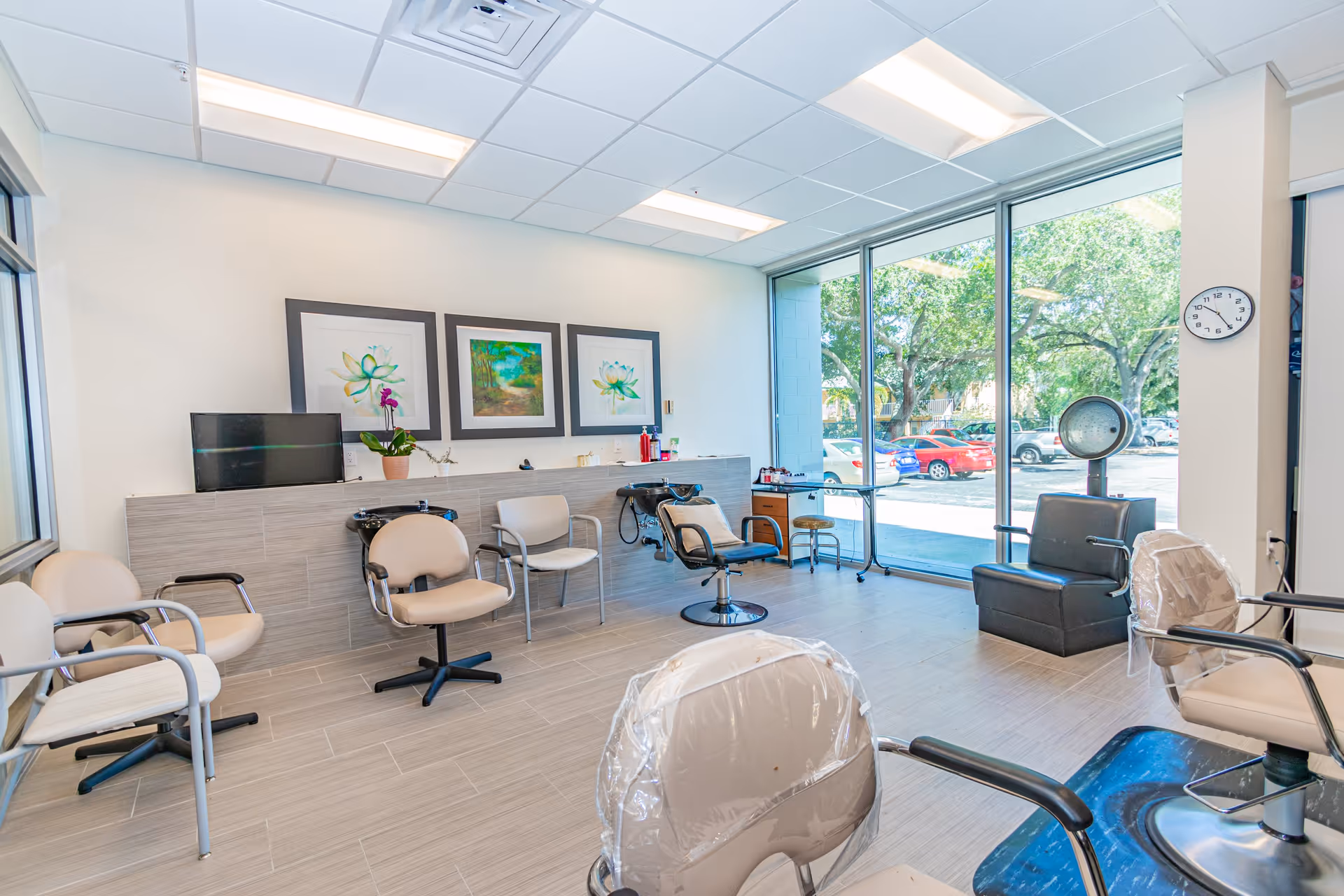 Bright and clean hair salon area with several beige salon chairs, two black shampoo stations, a black hair dryer chair, large windows showing a parking lot and trees outside, and three framed artworks on the wall.