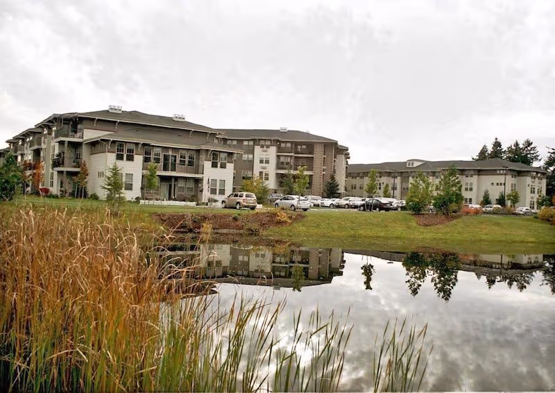 Exterior view of a senior living facility named Spring Creek by Bonaventure, showing a multi-story building with balconies and windows, a parking lot with several cars, green lawn, and a pond in the foreground reflecting the building and surrounding trees.