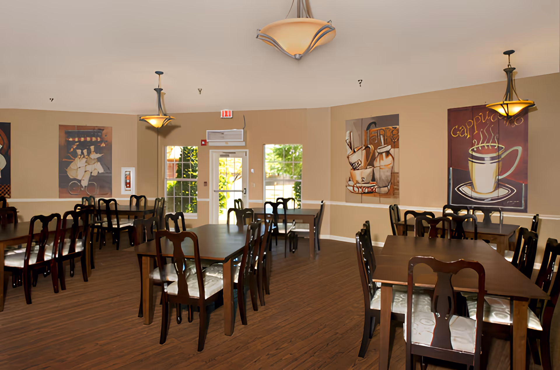 A dining room with multiple wooden tables and chairs arranged neatly. The room has beige walls adorned with large paintings, including one of a steaming cup labeled 'Cappuccino'. There are three hanging light fixtures providing warm lighting, and windows and a door letting in natural light.