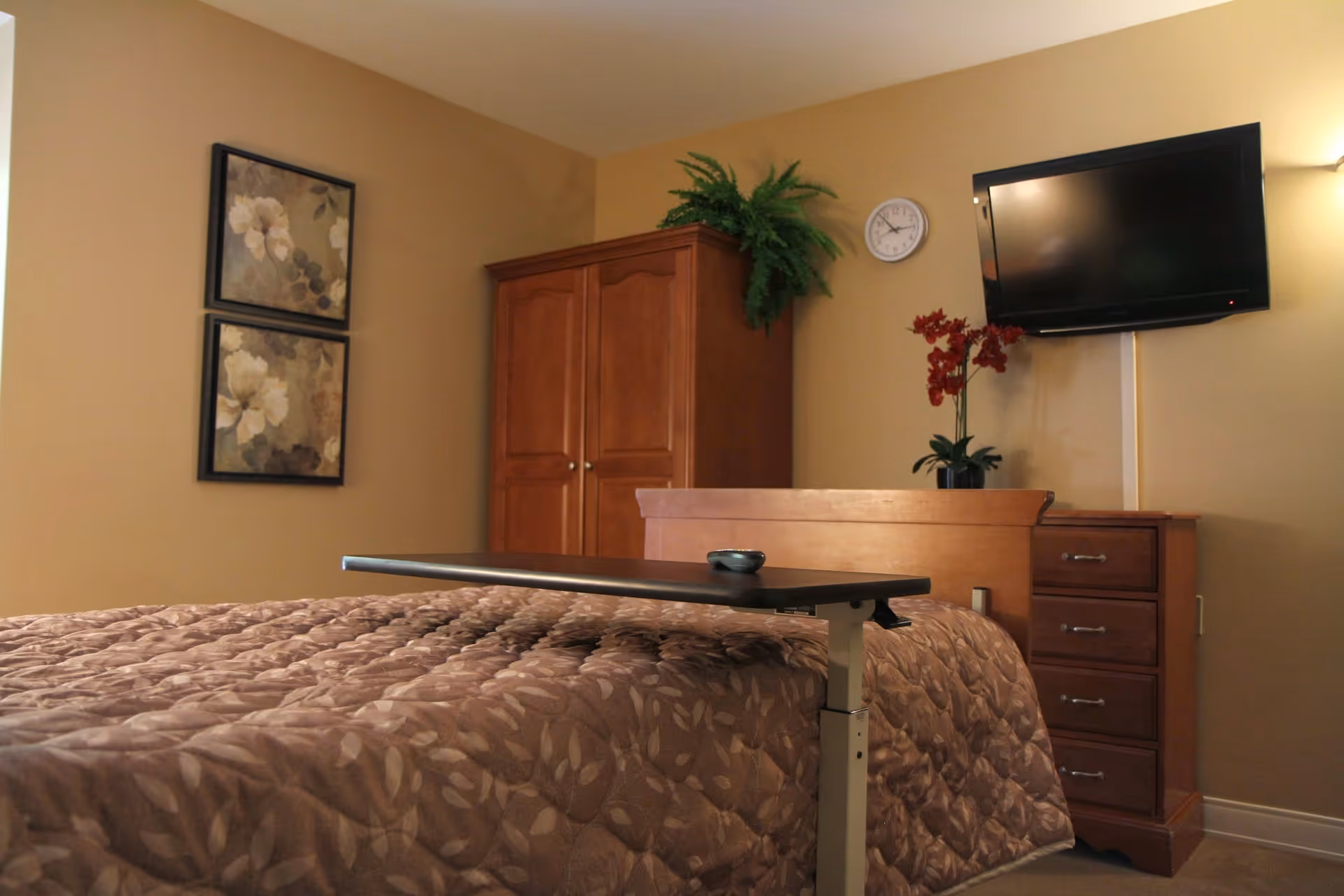 A bedroom with a bed covered in a brown patterned quilt, a wooden headboard, and a small adjustable table on the bed. On the wall above the headboard is a flat-screen TV and a round clock. There is a wooden dresser with drawers next to the bed, decorated with a plant with red flowers. A wooden wardrobe with a green plant on top is in the corner. Two framed floral paintings hang on the adjacent wall.