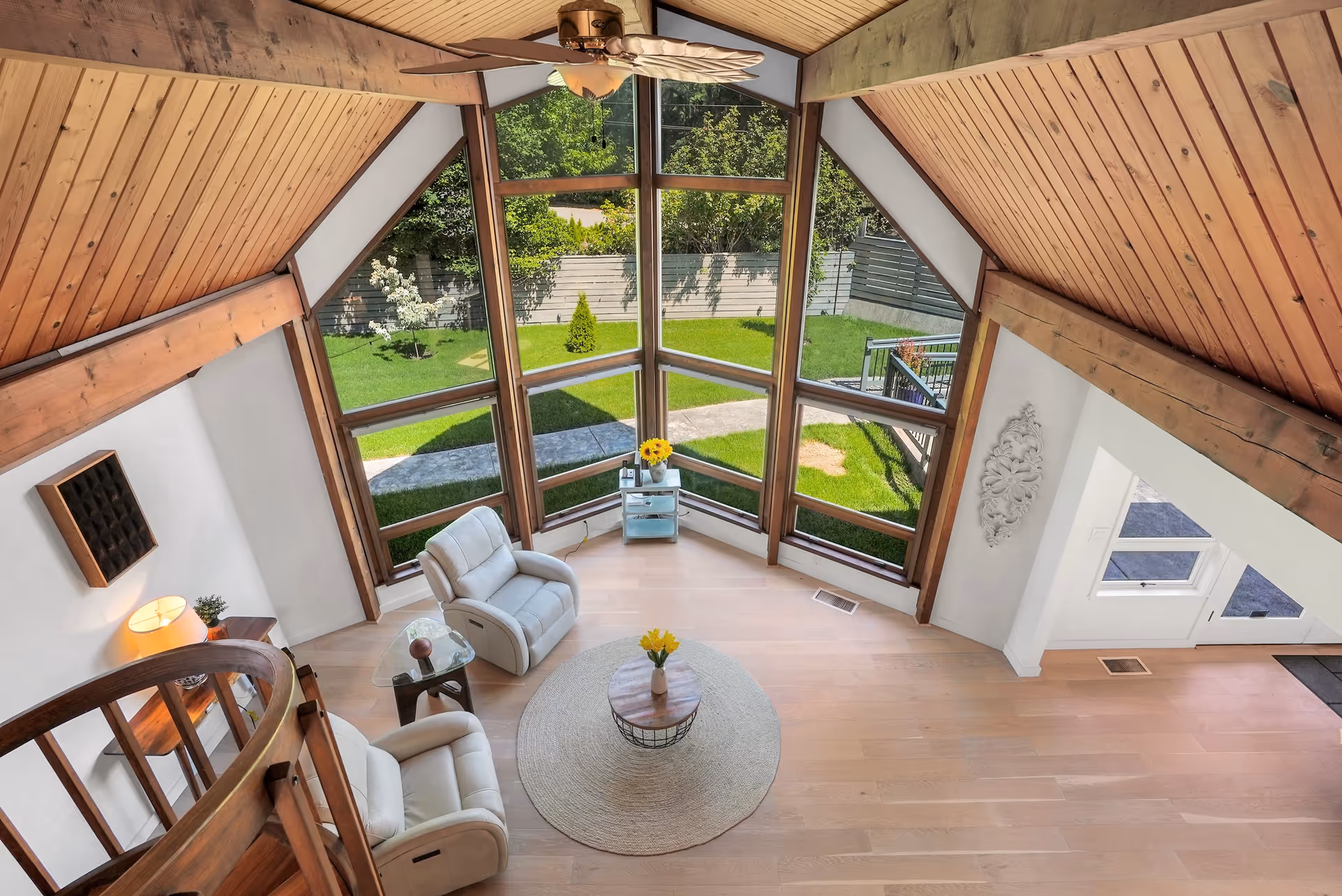A bright living room with large triangular windows overlooking a green garden. The room features two white recliner chairs, a round rug with a small coffee table holding a vase of yellow flowers, a wooden spiral staircase, and a ceiling fan with wooden blades. The ceiling and beams are made of wood, creating a warm and cozy atmosphere.