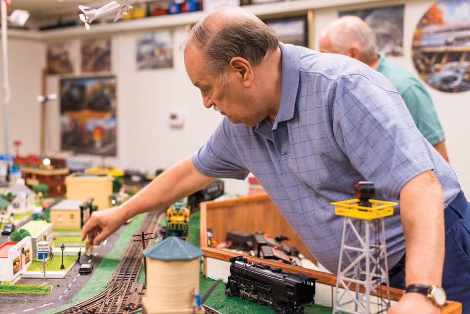 An elderly man in a blue checkered shirt is leaning over a detailed model train set, adjusting a small figure on the miniature road. Another elderly man is visible in the background. The room has various model train-related decorations and pictures on the walls.