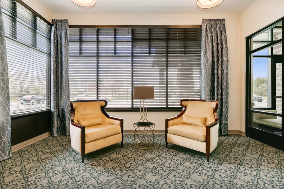 A bright seating area with two upholstered armchairs facing each other, a small side table and lamp between them, and large windows with blinds and patterned curtains.