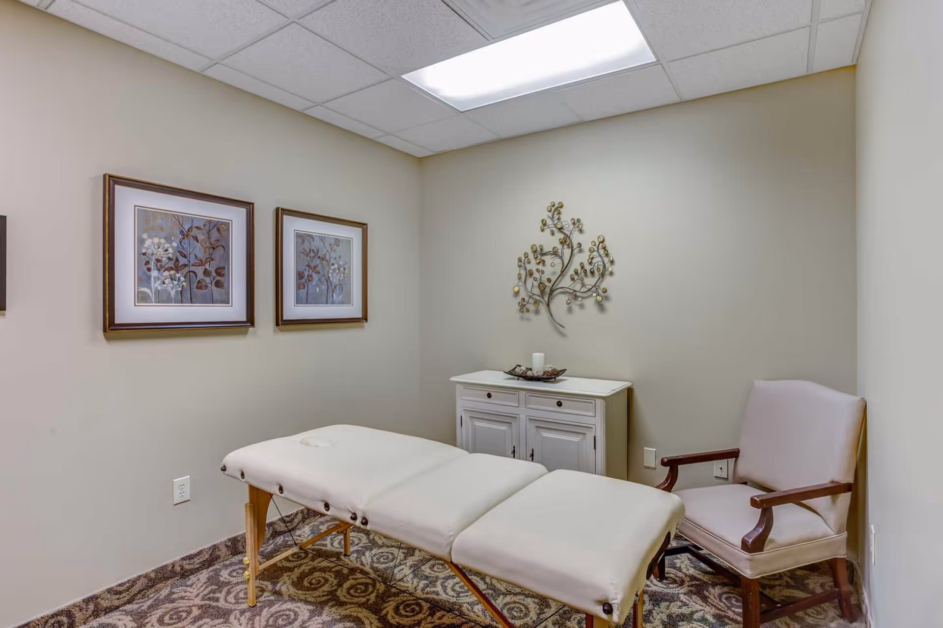 Small treatment room with a padded massage table, armchair, decorative cabinet and wall art.