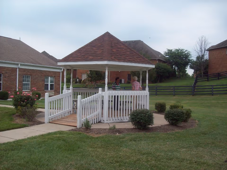 Two people standing and talking inside a white wooden gazebo with a shingled roof, located on a grassy lawn with bushes and surrounded by brick buildings and a black wooden fence.