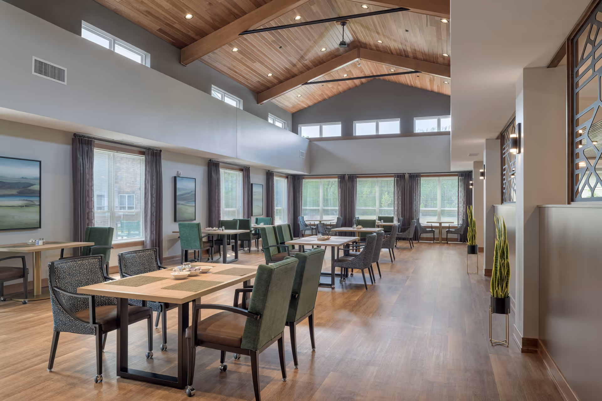 A spacious and well-lit dining room in a senior living facility with wooden floors, high wooden ceiling with beams, multiple tables and chairs arranged neatly, large windows with curtains allowing natural light, and decorative plants along the walls.