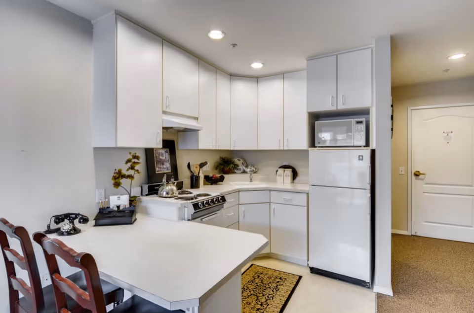 A compact kitchen area with white cabinets and countertops, featuring a stove with a kettle, a microwave above a refrigerator, and a small dining counter with two wooden chairs. The kitchen is well-lit with recessed ceiling lights and has a decorative rug on the floor. A hallway with a closed white door is visible in the background.