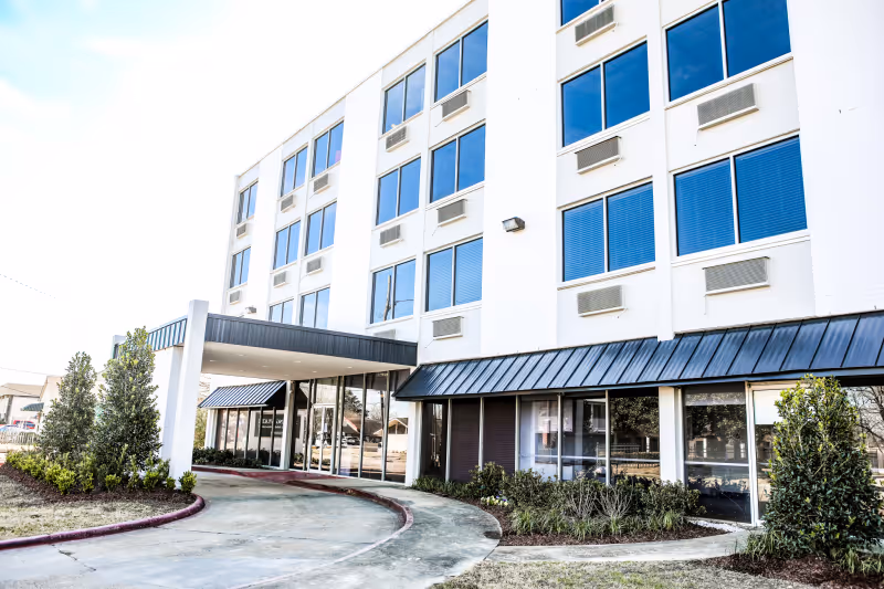 Front entrance and facade of a multi-story white building with a covered porte-cochere, large windows, and landscaping.