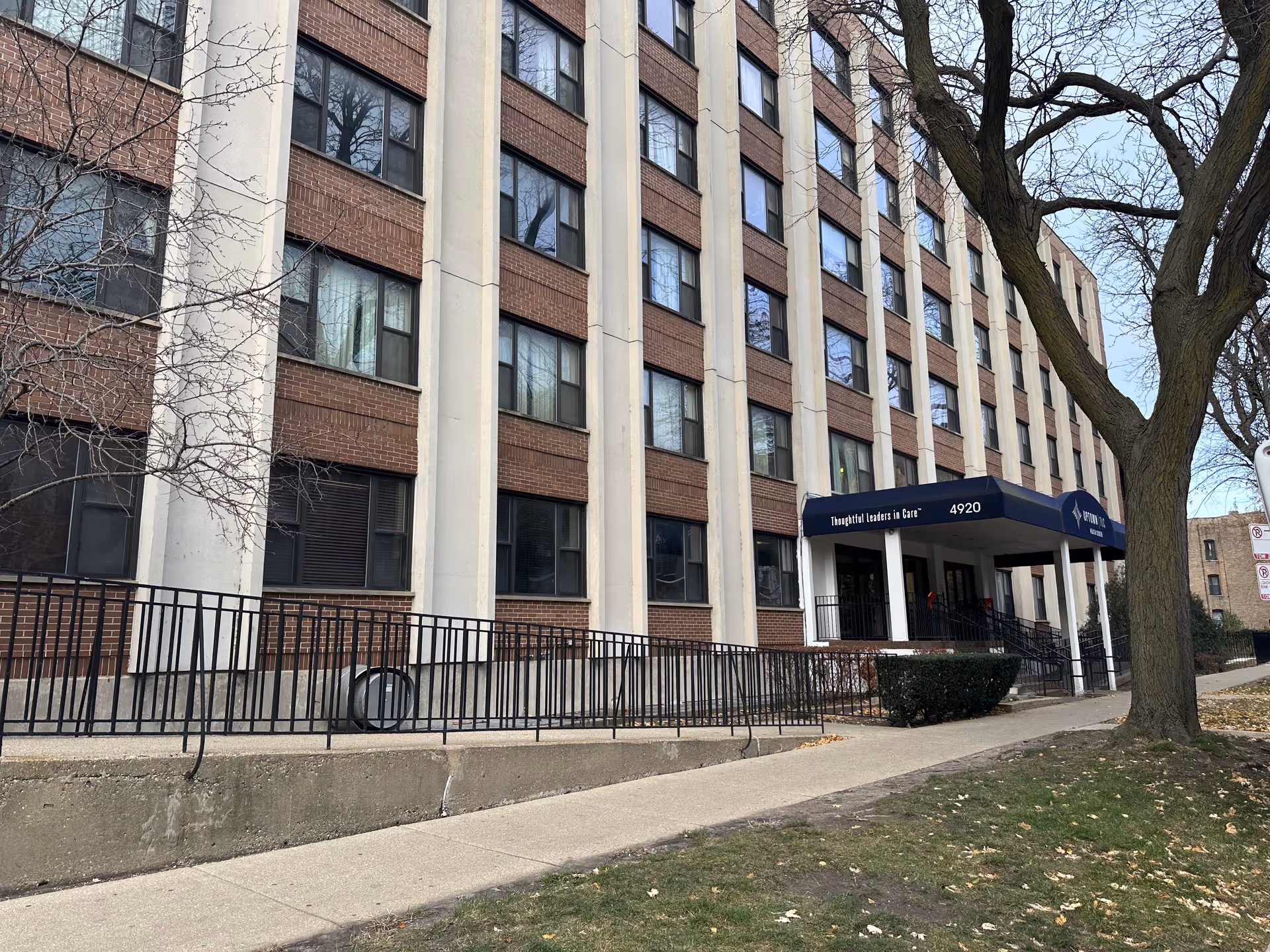 Front exterior of a multi-story brick care building with a blue awning over the entrance and a sidewalk.