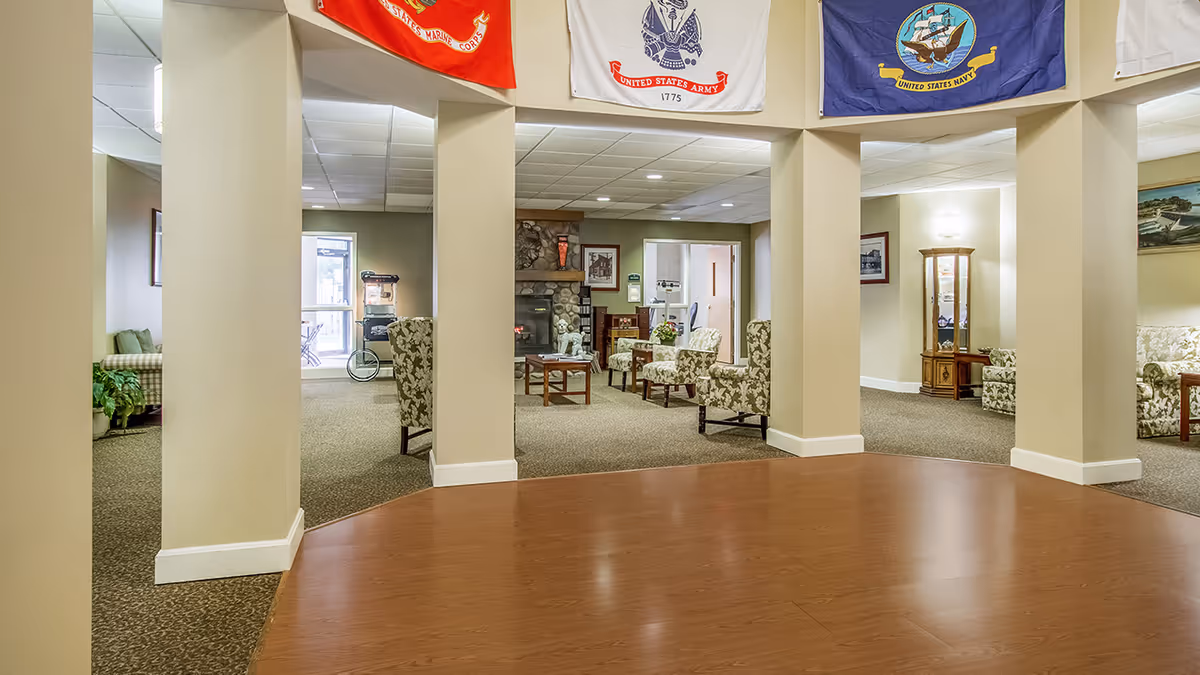 Interior view of a senior living community common area with beige walls and columns, carpeted seating areas with floral upholstered chairs, a stone fireplace, and flags of the United States Marine Corps, Army, and Navy hanging from the ceiling.